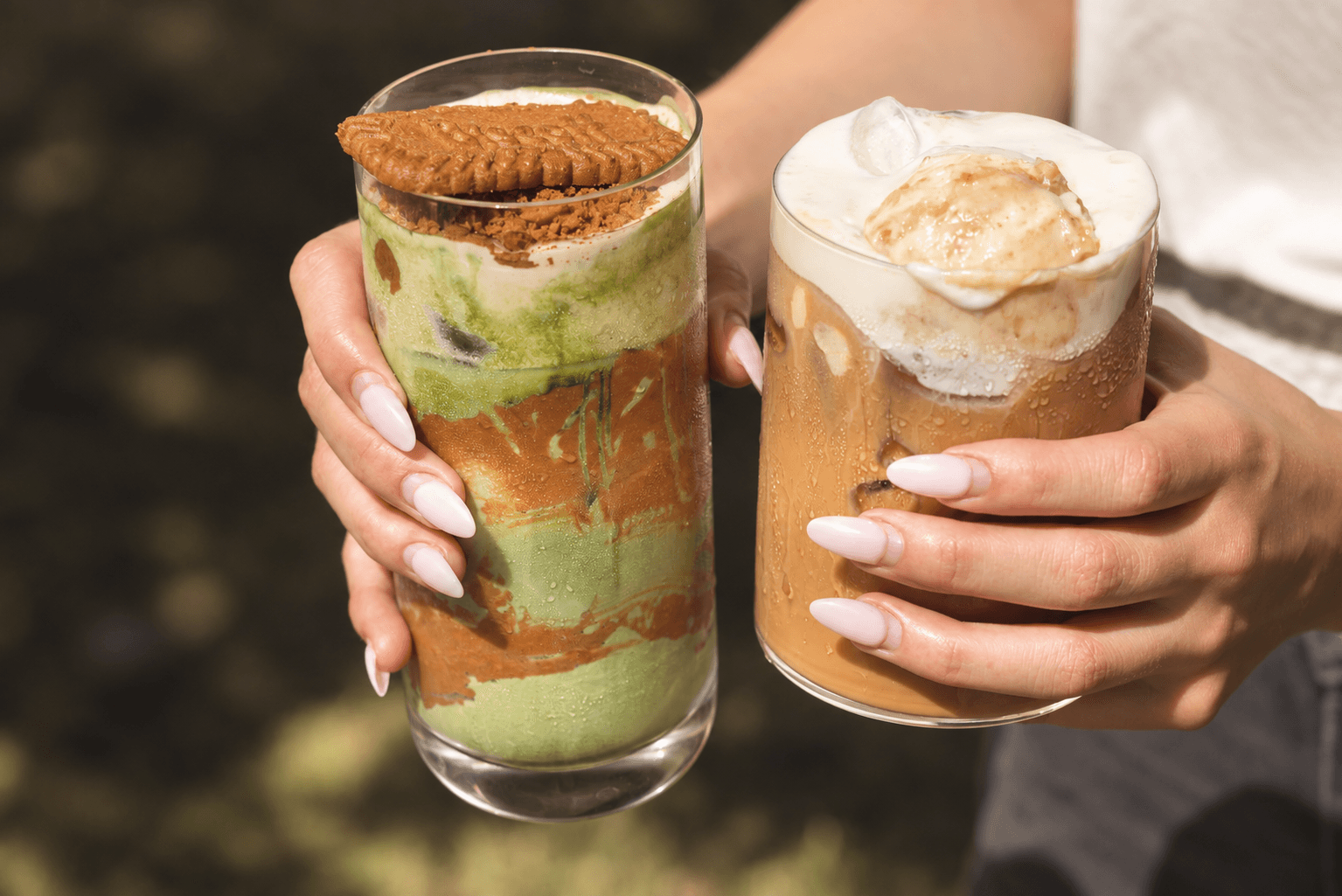 Two Cafe Mallow drinks held in the sunlight: a layered matcha with a Biscoff cookie and a creamy iced coffee.